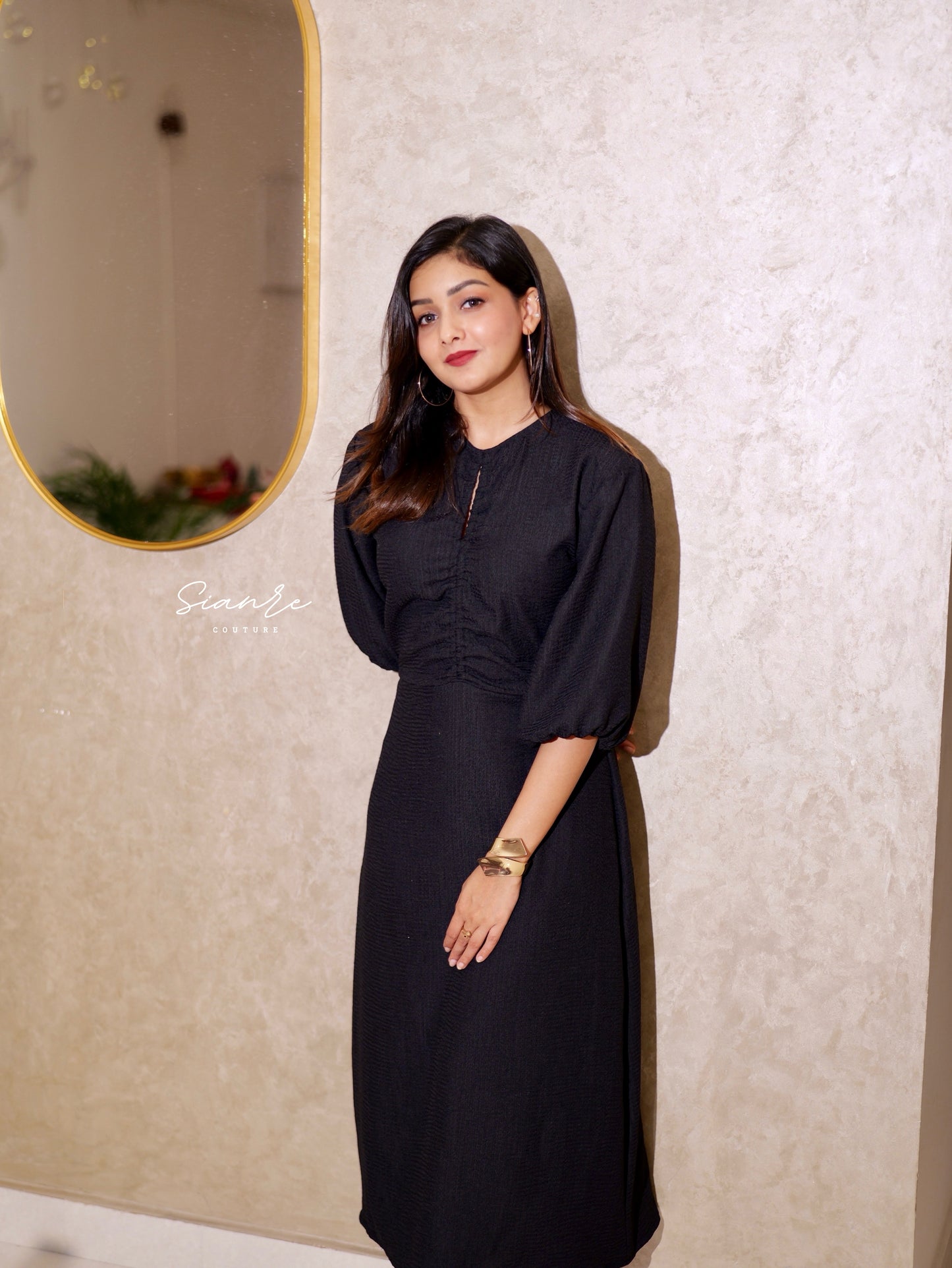 Woman in a black dress standing against a textured wall with a mirror.