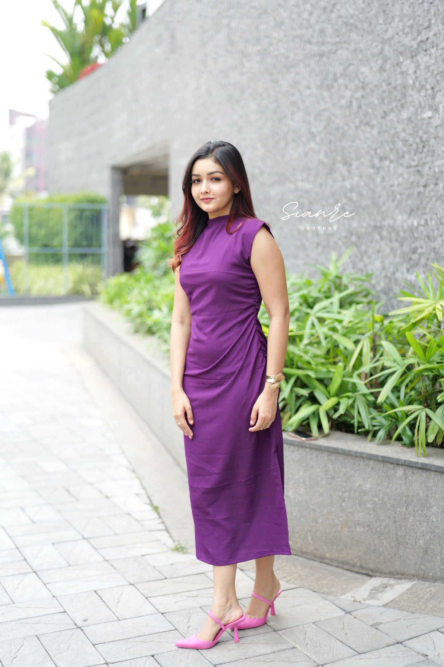 Woman in a purple dress standing outdoors with greenery and a building in the background