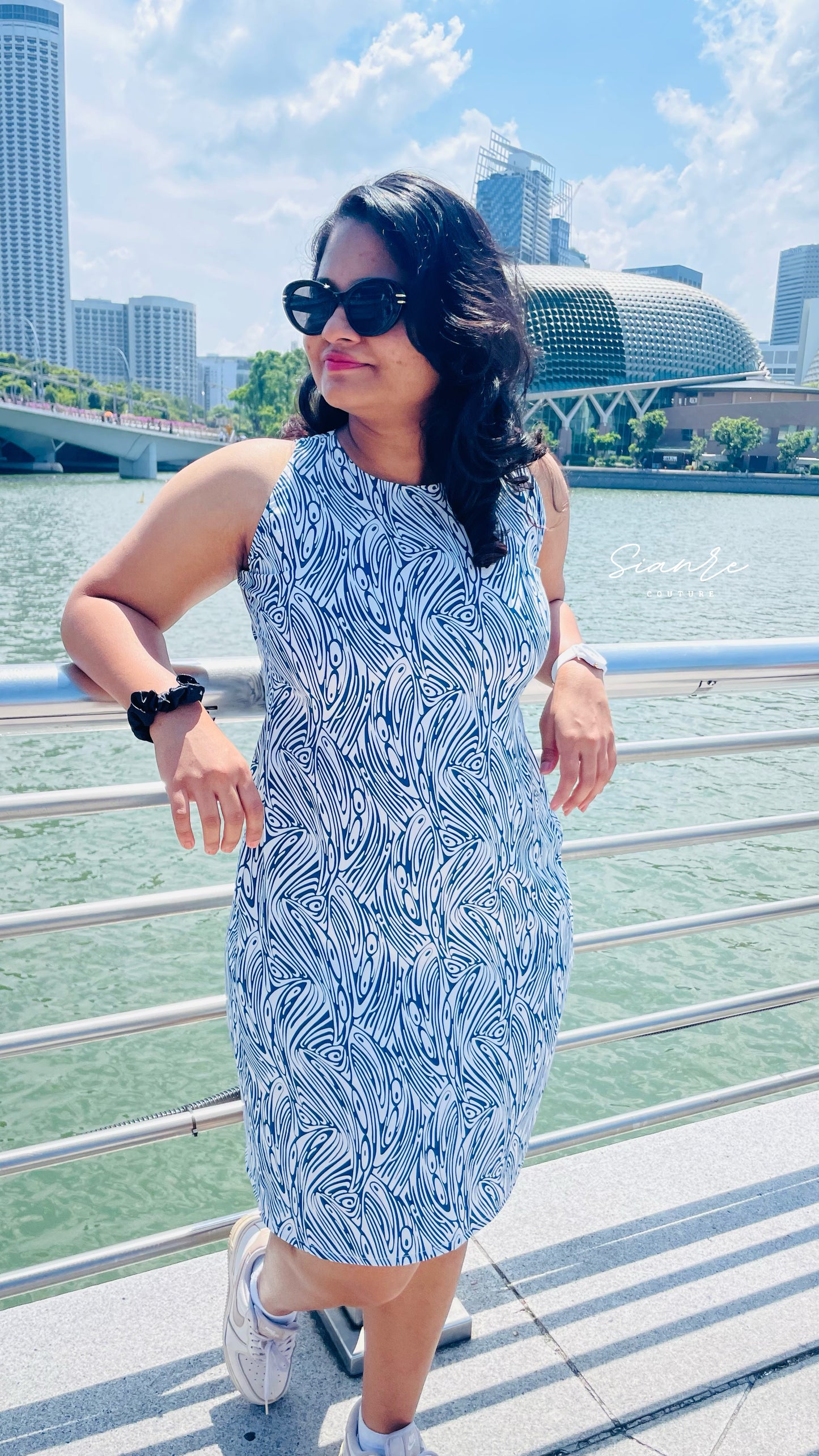Woman in a blue patterned dress standing by a waterfront with city skyline in the background