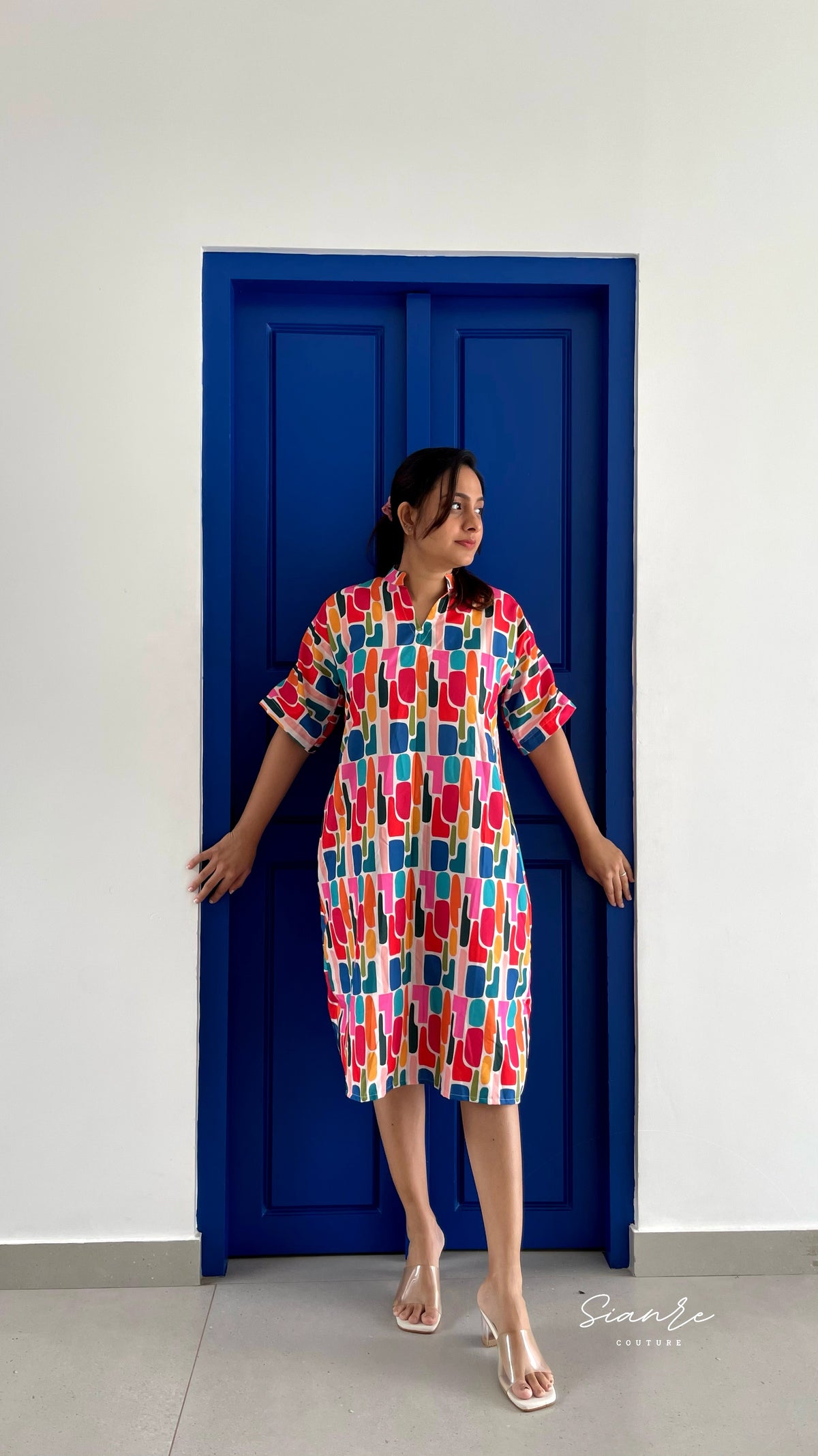 A woman wearing a colorful, short-sleeved midi dress with an abstract geometric pattern in red, blue, yellow, and white, posing in front of a vibrant blue door on a white wall.