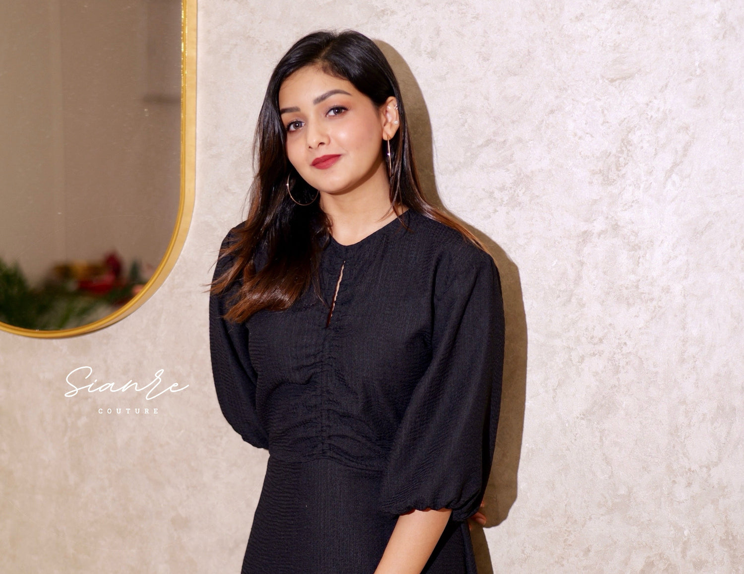 Woman in a black dress standing against a textured wall with a mirror.