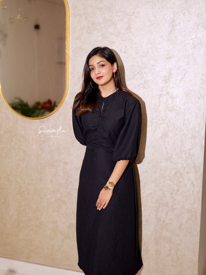 Woman in a black dress standing against a textured wall with a mirror.