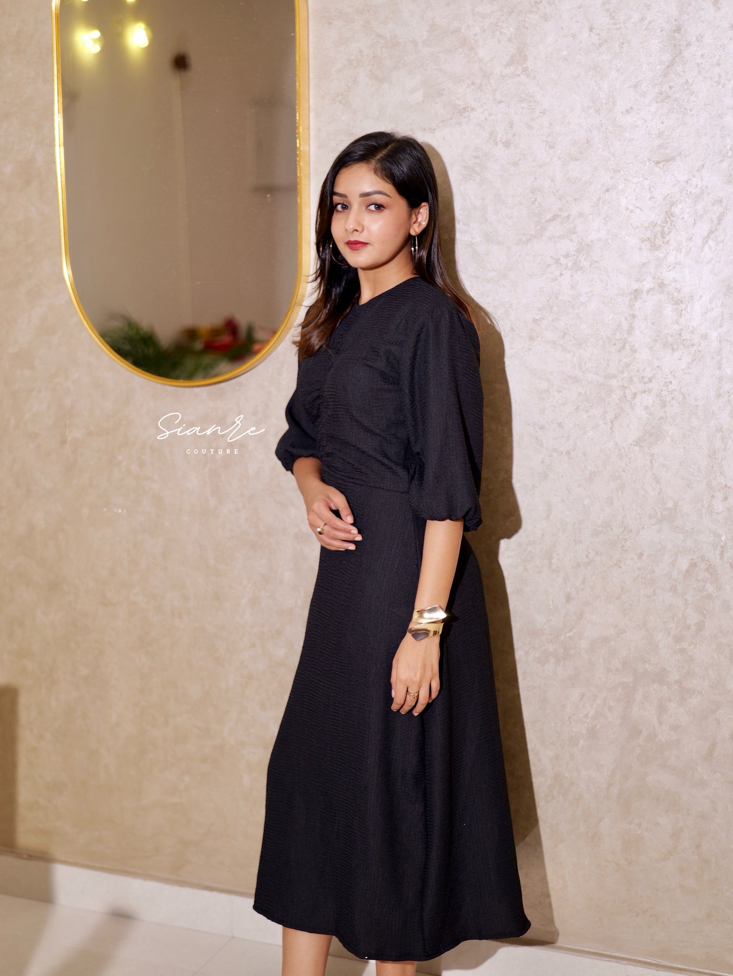 Woman in a black dress standing against a textured wall with a mirror.