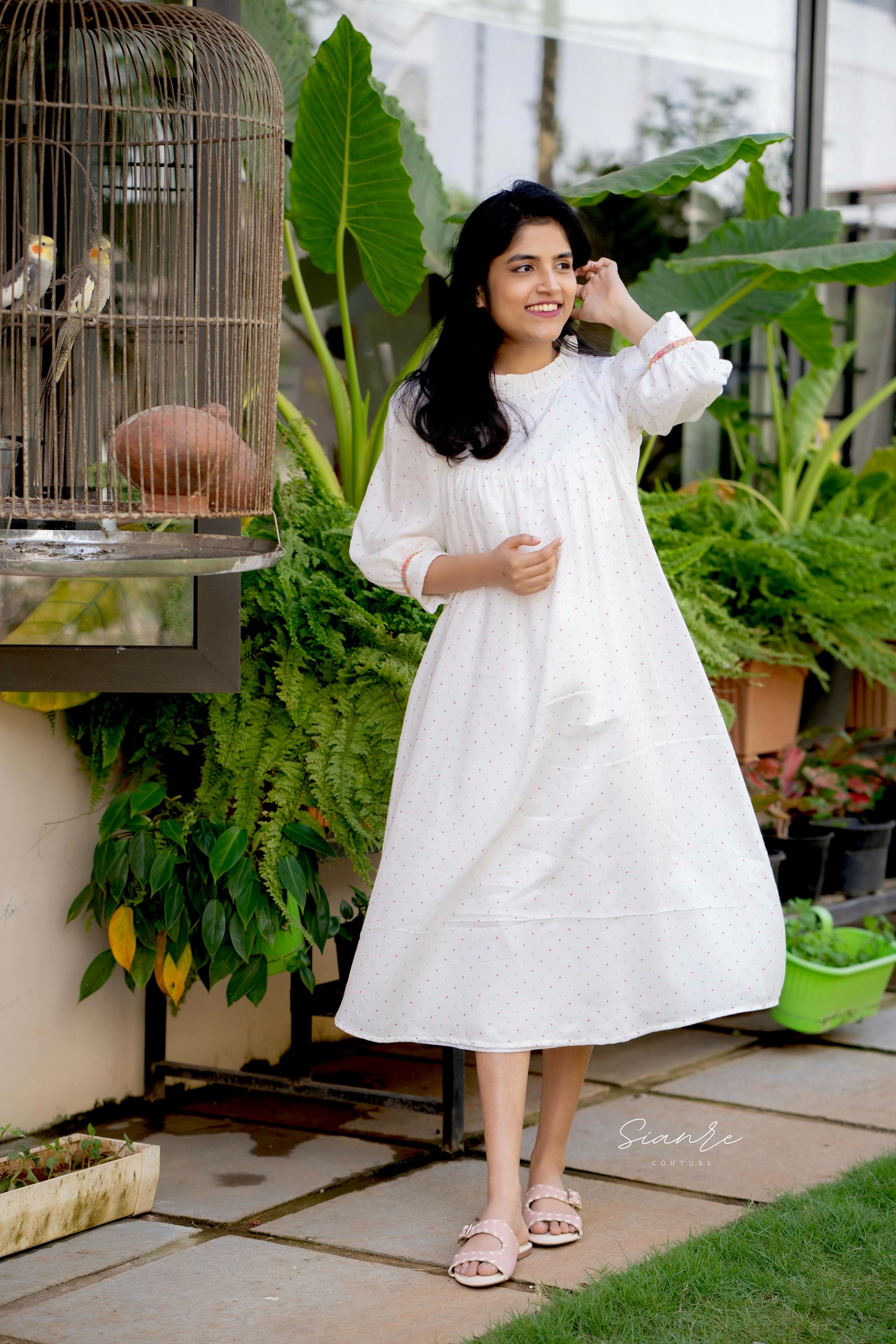 Woman in a white dress standing in a garden with plants and a birdcage.