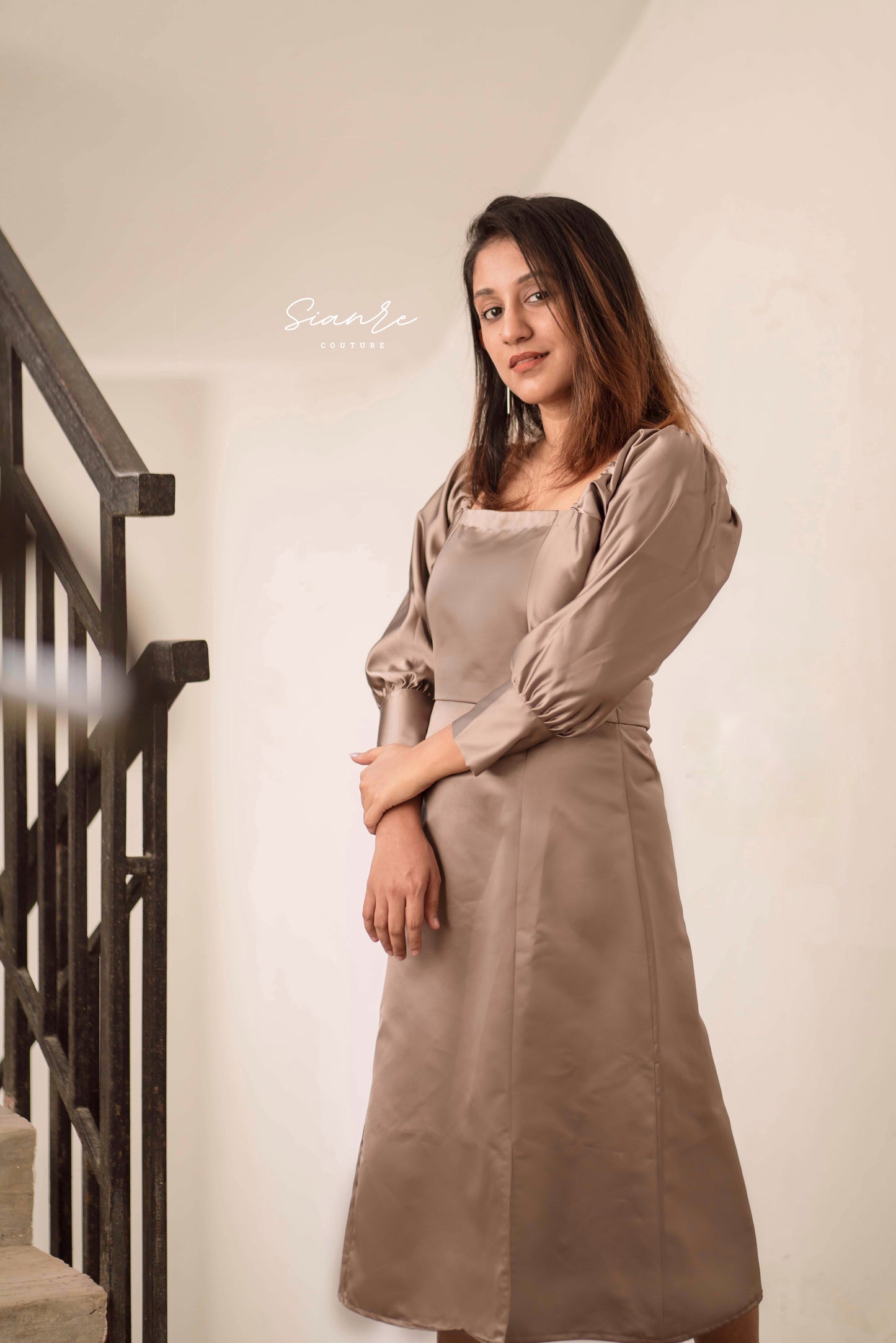 Woman wearing a beige dress standing against a white wall with a staircase.