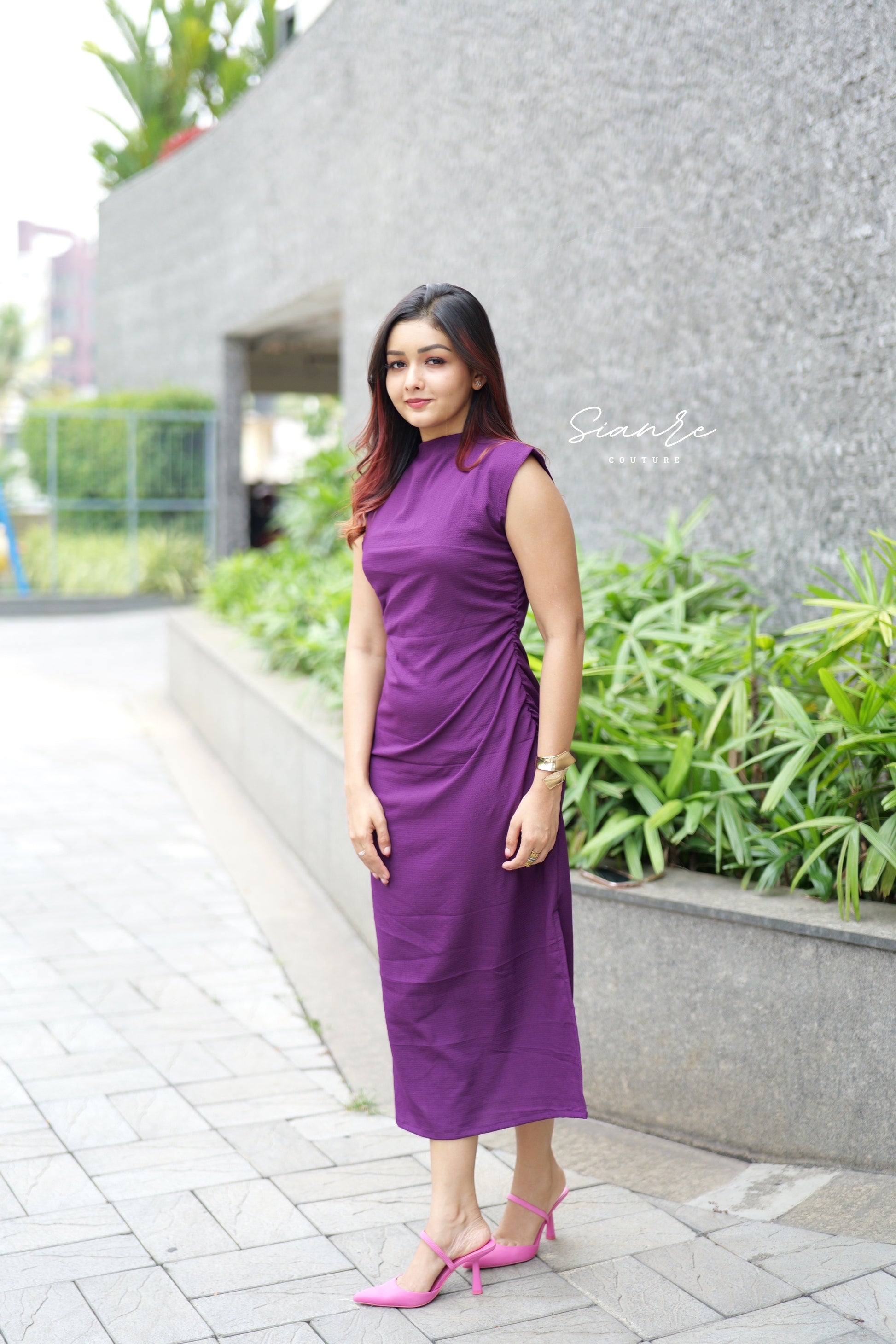 Woman in a purple dress standing outdoors with greenery and a building in the background