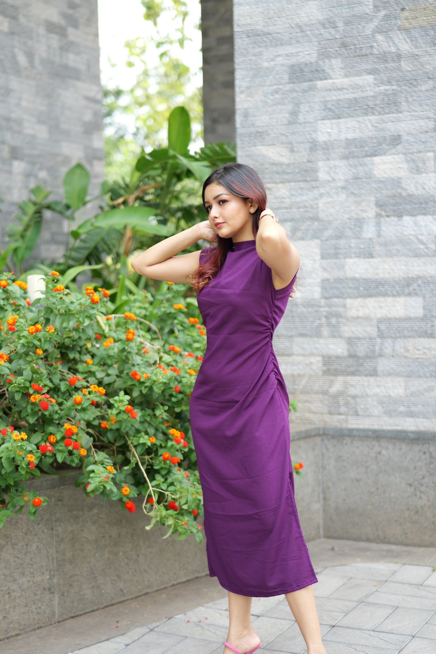 Woman in a purple dress standing outdoors with a gray wall and plants in the background