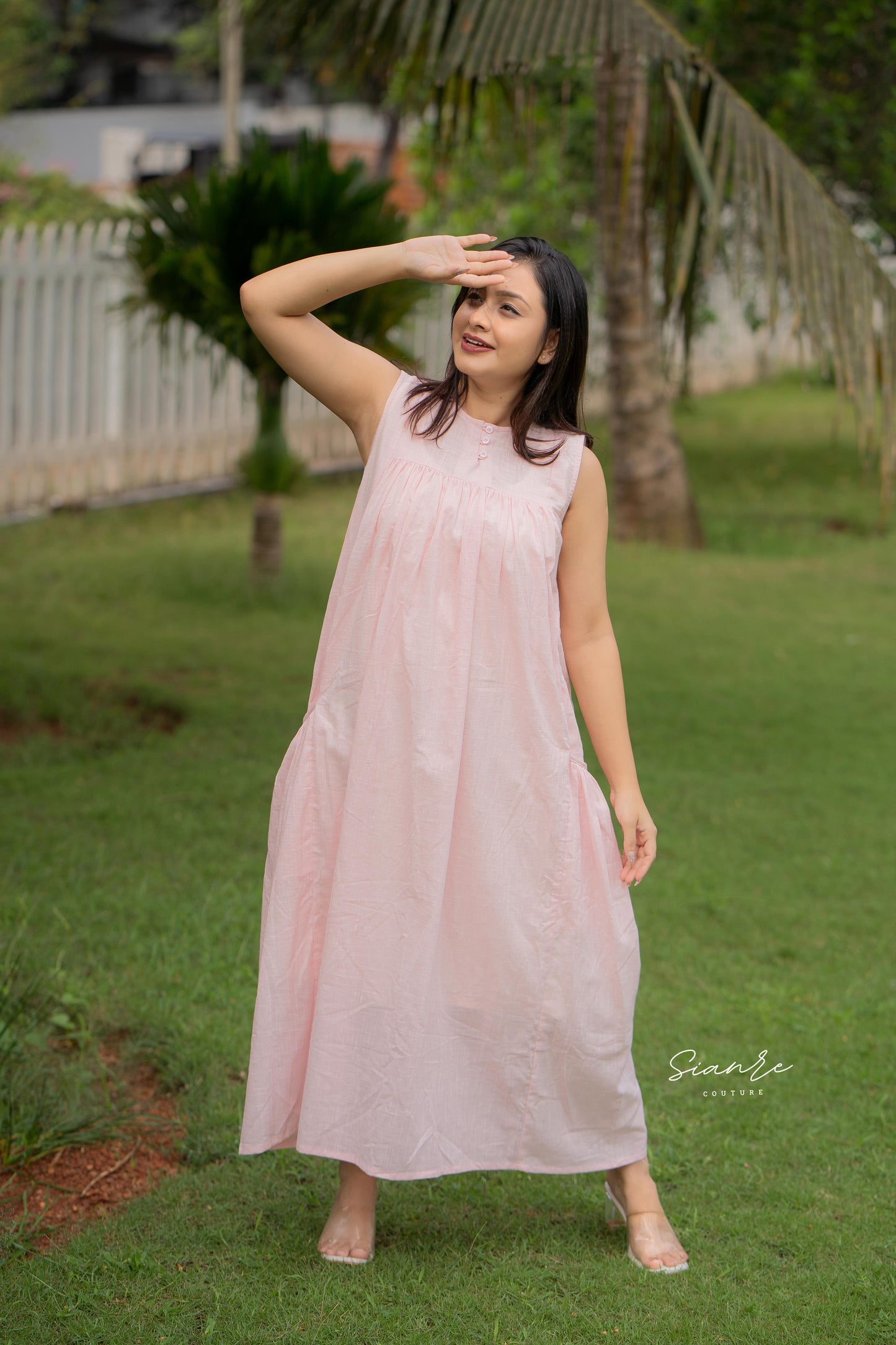 Woman in a light pink sleeveless dress standing on grass with trees in the background